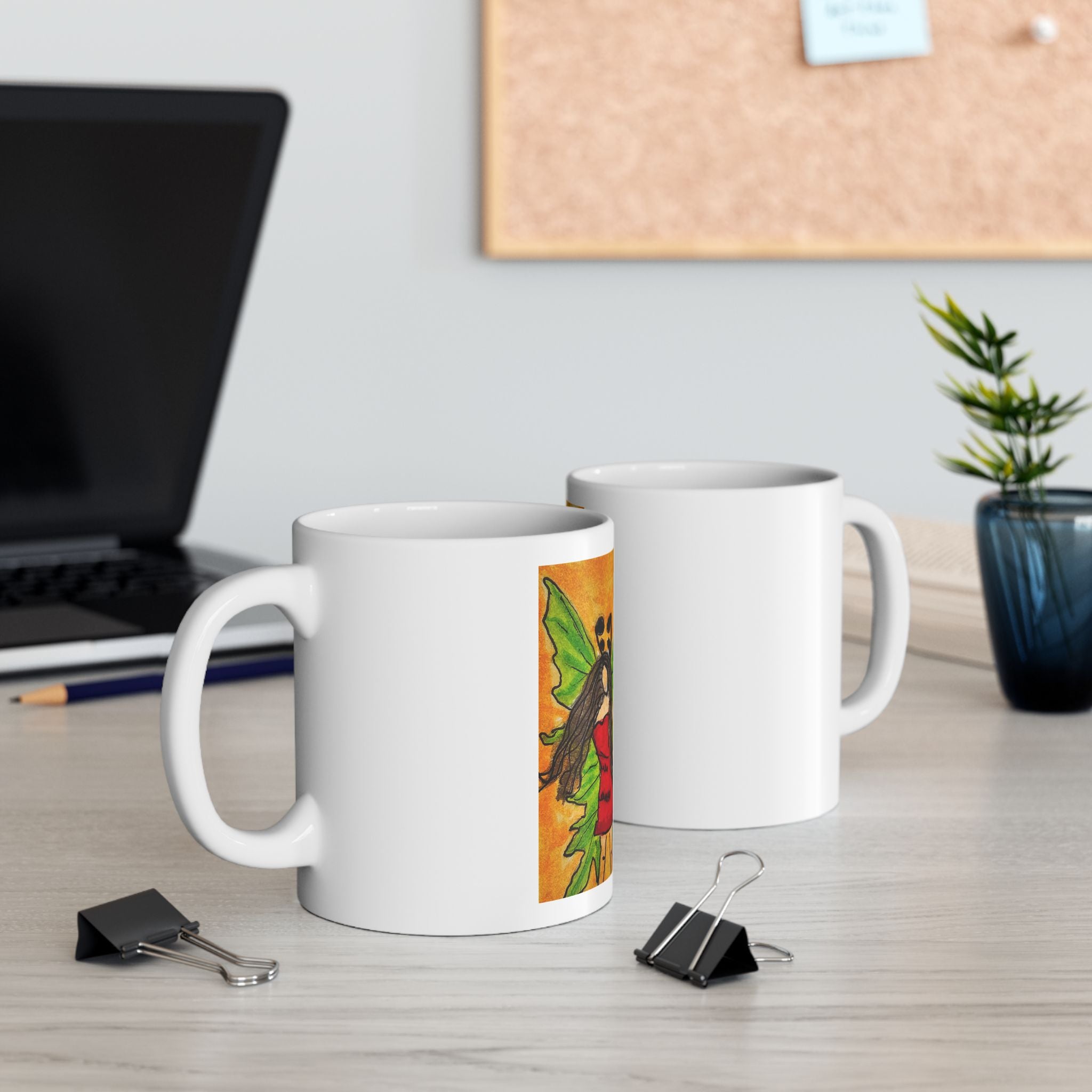 White ceramic mugs on a desk, each with the green-winged fairy artwork on the front.