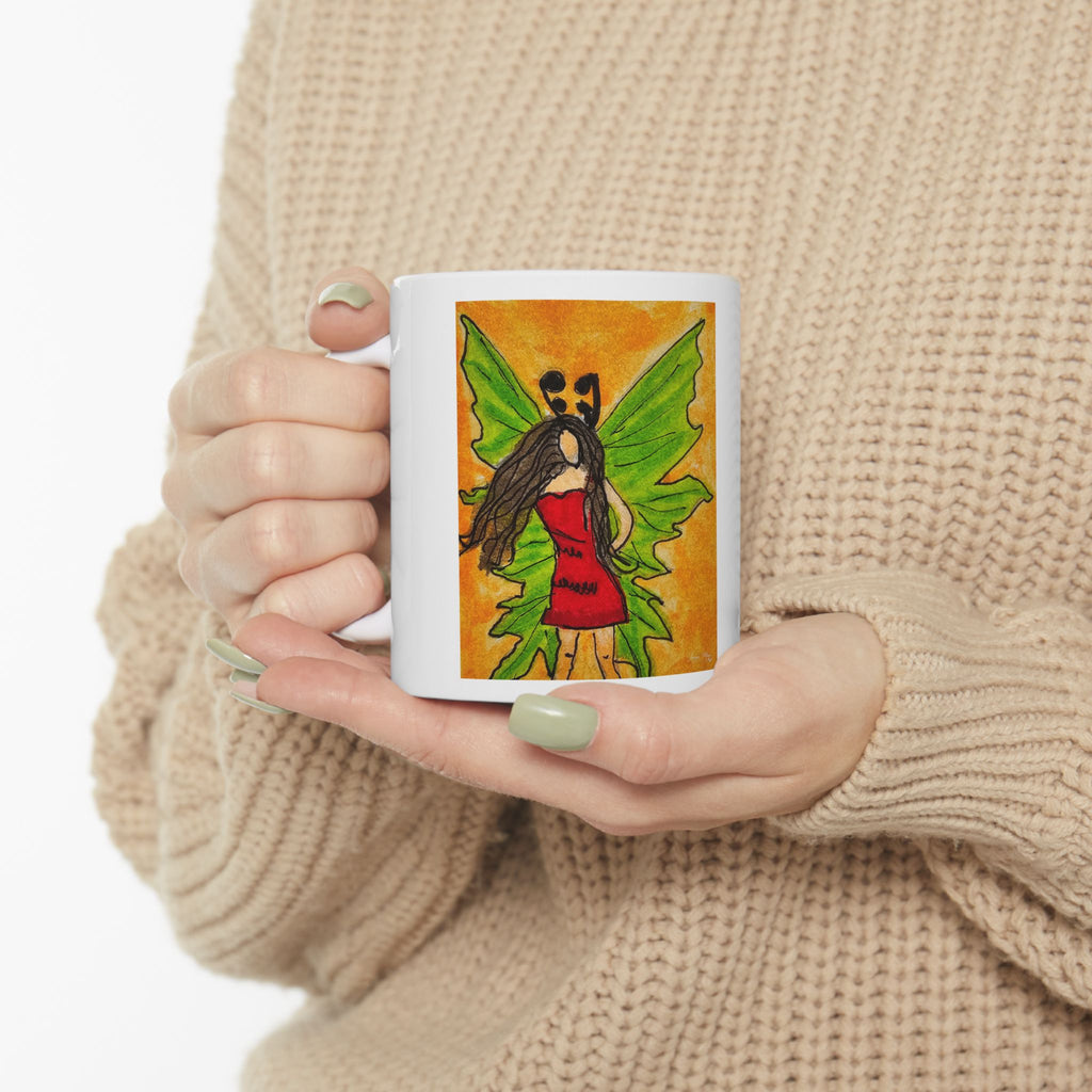 White ceramic mug held by hands, featuring a green-winged fairy illustration on the front.