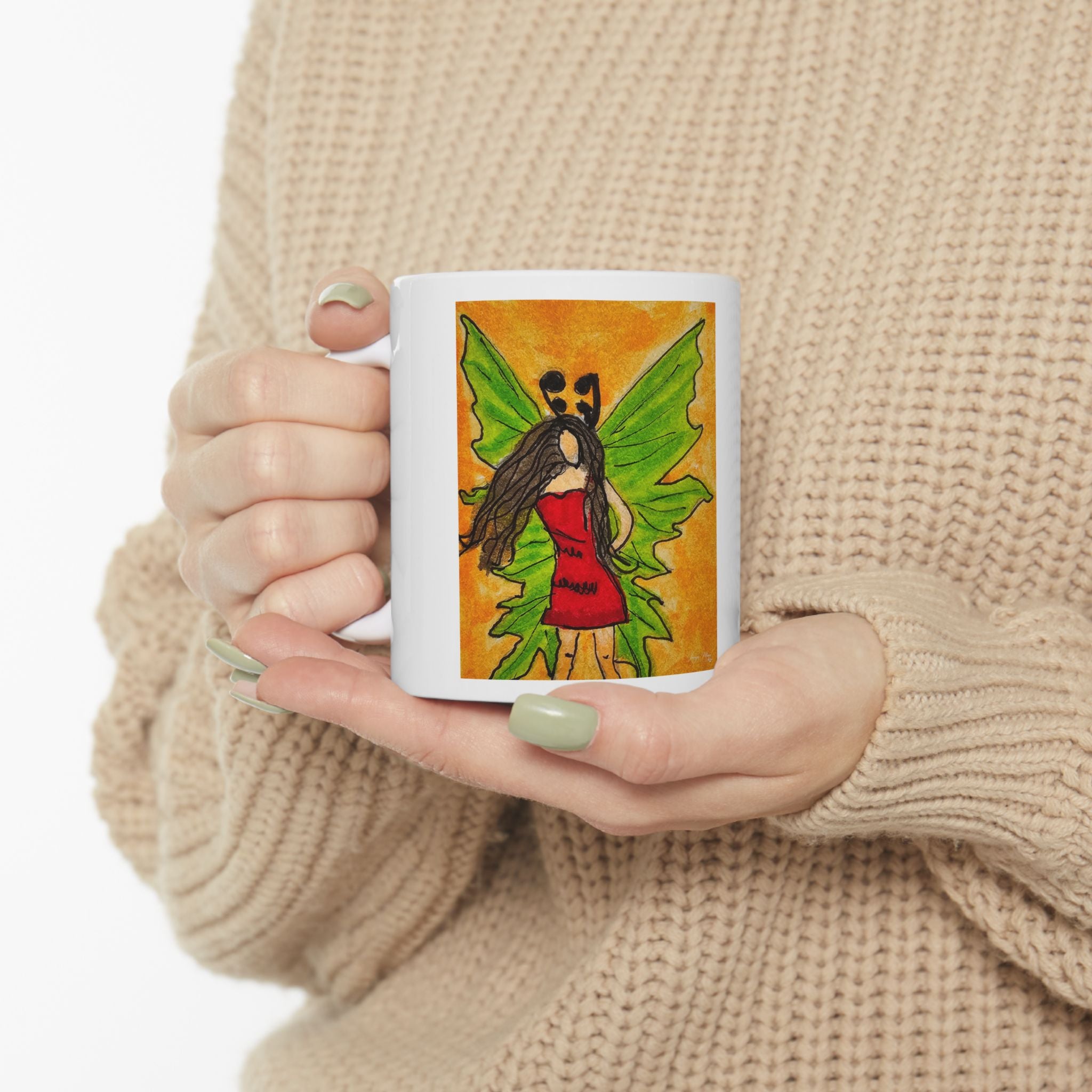 White ceramic mug held by hands, featuring a green-winged fairy illustration on the front.