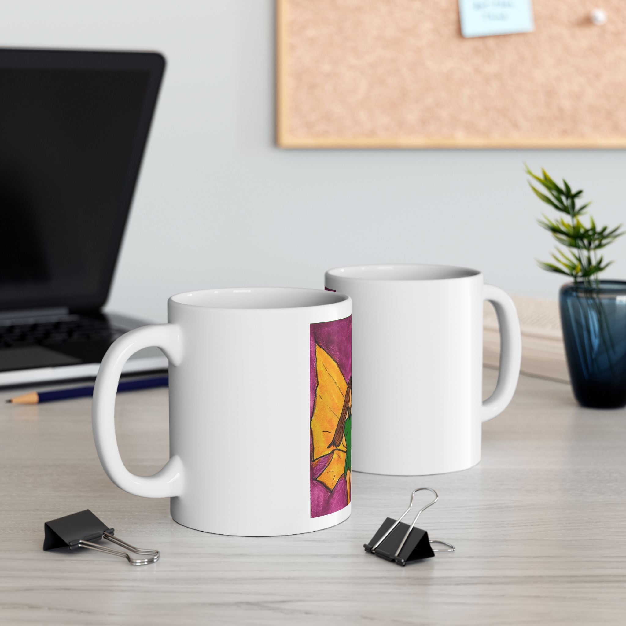 Two white ceramic mugs on a desk, one showing a colorful fairy wing watercolor panel.